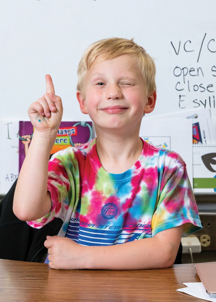 Image of child smiling at camera and pointing in classroom setting.